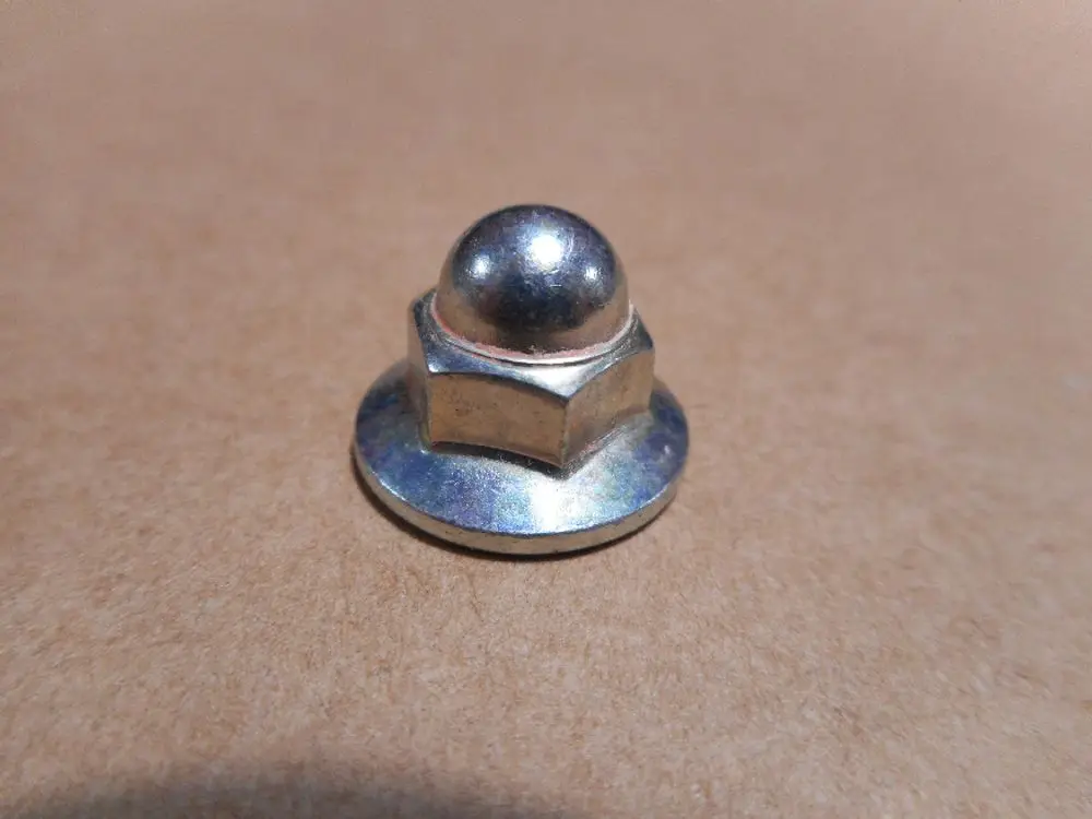 Mutter aus Metall mit einer gewölbten Oberseite und einer sechseckigen Basis, auf einem beige Hintergrund platziert. Herstellerangabe: Remeha.